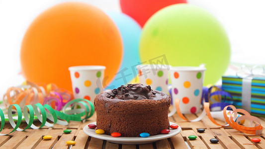 质朴的木桌上的巧克力生日蛋糕，背景是彩色气球、礼物、塑料杯和糖果，背景是白墙