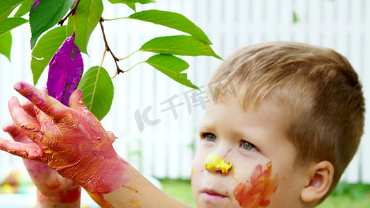 一个小孩，一个四岁的男孩在夏天玩耍，用手指画画，在花园里的树上装饰树叶。