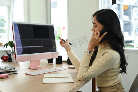 年轻的女性视频编辑坐在人事电脑前的创意工作场所，用手机聊天