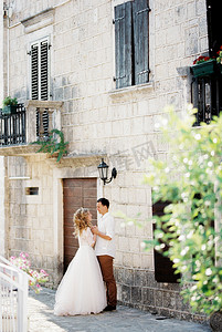 新郎握着新娘的手站在旧石屋附近，窗户上有百叶窗
