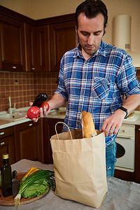 物品摆好摄影照片_英俊的白人男子在厨房里摆好采购的物品，在打开杂货购物袋的同时分拣蔬菜