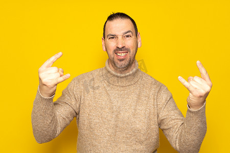 穿着米色高领毛衣的兴奋时髦男人的照片在孤立的黄色背景上展示了两个沉重的摇滚手势。