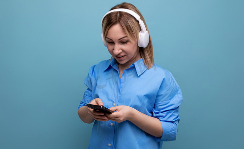 一位身穿休闲衬衫的千禧一代女性的特写，她戴着白色大耳机，在蓝色孤立背景中专注于音乐，有复制空间