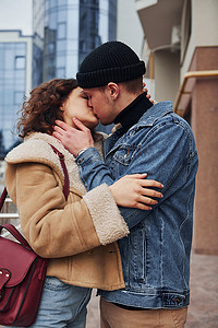 在商业大楼附近的城市里，一对穿着休闲保暖衣服的快乐情侣在户外接吻