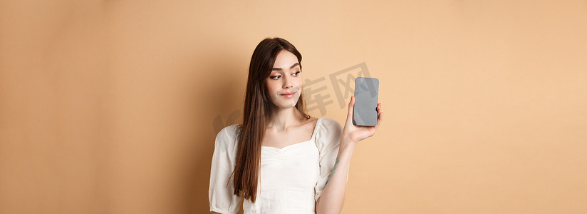 站在米色背景中，迷人的年轻女性展示着空荡荡的智能手机屏幕，微笑着望向一边