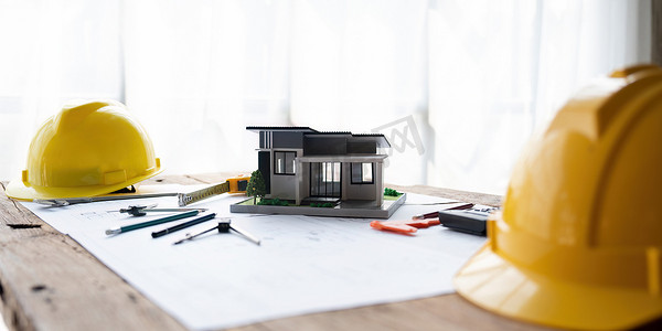 办公桌上房屋的建筑模型，带有绘图技术工具安全头盔和建筑施工计划、室内设计师和建筑师工作概念的蓝图卷