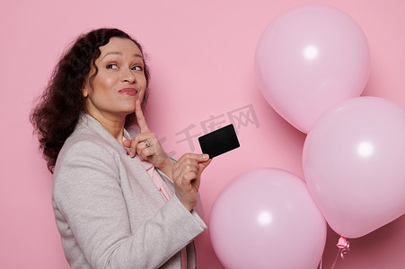 粉色梦幻背景摄影照片_一个快乐的梦幻女人的肖像拿着借记卡和智能手机，孤立的粉红色背景。