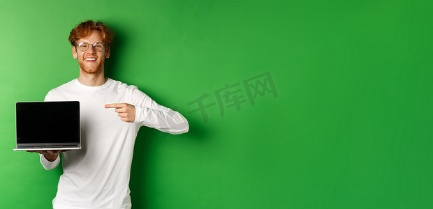 休闲长袖摄影照片_戴着眼镜和白色长袖 T 恤的快乐红头发人，手指指着空白的笔记本电脑屏幕，微笑着，站在绿色背景上