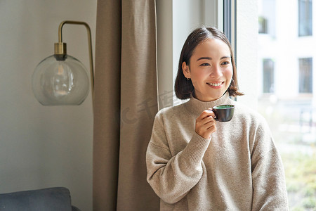 美丽的亚洲女人坐在家里喝杯咖啡，一边享用浓缩咖啡，一边看着窗外的路人，开心地笑着