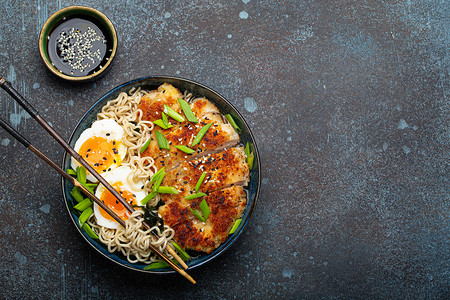 亚洲面条拉面汤、油炸的 panko 鸡片和煮鸡蛋在陶瓷碗里用筷子和酱油在石头质朴的背景顶视图上，文字空间