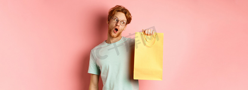 红头发、戴着眼镜和 T 恤、拿着并看着黄色购物袋、在促销活动中购买礼物、站在粉红色背景上的风趣帅哥