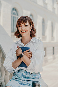 一位身穿白衬衫和蓝色牛仔裙的女士坐在栏杆上，将蓝色记事本抱在建筑物上。