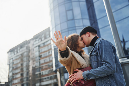 保暖衣服摄影照片_在商业大楼附近的城市里，一对穿着休闲保暖衣服的快乐情侣在户外接吻