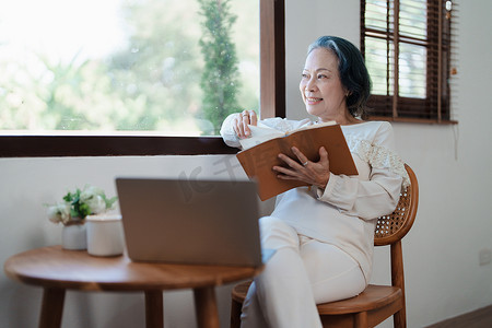一位亚洲老年妇女的画像，摆出现代姿势，手持记忆笔记本，操作电脑。