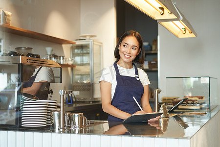 美丽的亚洲女孩微笑的肖像，咖啡师在柜台后面工作的咖啡馆，使用平板电脑作为 POS 终端，处理订单