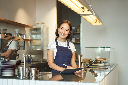 美丽的亚洲女孩微笑的肖像，咖啡师在柜台后面工作的咖啡馆，使用平板电脑作为 POS 终端，处理订单