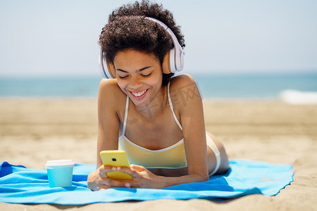 戴着耳机在海边浏览智能手机的快乐民族女游客