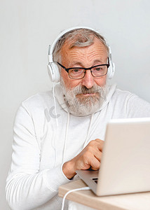 身着连帽衫和眼镜的肖像特写帅老头正在使用笔记本电脑，一边坐在家里的沙发上一边微笑着听音乐