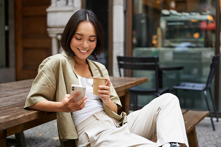 时尚的都市女孩坐在咖啡馆里，拿着手机喝咖啡聊天，在智能手机上浏览社交媒体