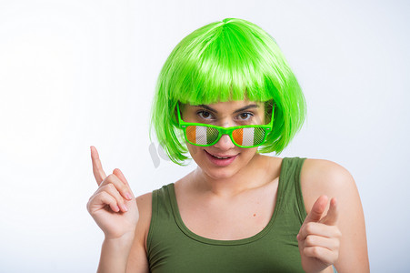 假发摄影照片_戴着绿色假发和滑稽眼镜的快乐年轻女性在白色背景下庆祝圣帕特里克节