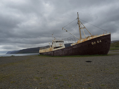 冰岛，西峡湾，Patreksfjordur，2018 年 6 月 24 日：旧的废弃生锈渔船沉船站在冰岛西峡湾的沙滩海岸，戏剧性的灰色天空和悬崖背景