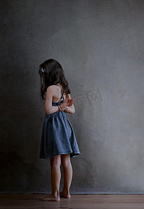 儿童理发摄影照片_受罚的女孩站在角落里，被整个世界冒犯了，一个身穿蓝色夏装、长着黑发的女孩的后视图，在深色背景下双手背在身后