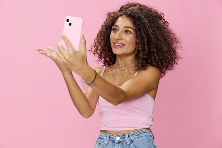 女博主手持电话视频通话，卷发粉色上衣，牛仔裤摆在粉色背景、复制空间、技术和社交媒体上，在线