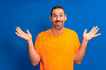 穿着橙色 T 恤、心满意足、无忧无虑、印象深刻的男人的肖像举起手掌，在蓝色背景中大喊“是”。