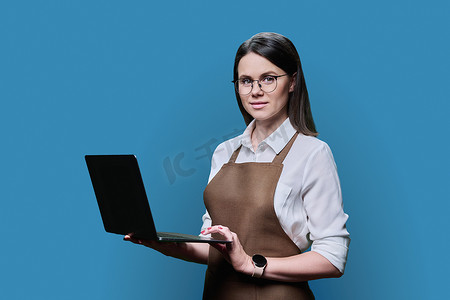 穿着围裙的年轻女性在蓝色工作室背景下使用笔记本电脑