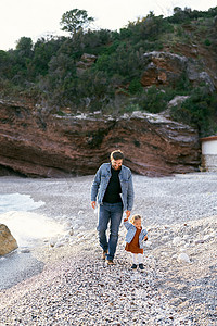 石子落下摄影照片_在绿色落基山脉的背景下，微笑的爸爸手牵着一个小女孩沿着鹅卵石海滩
