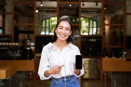 快乐的亚洲女人，咖啡馆经理站在餐厅前，指着智能手机应用程序，展示手机屏幕，推荐应用程序