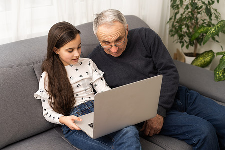 快乐的退休祖父和漂亮的孙女一边笑一边看电子书，通过笔记本电脑一起学习教育。
