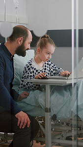 父亲和生病的女儿坐在医院病房里，一边用笔记本电脑玩在线治疗视频游戏