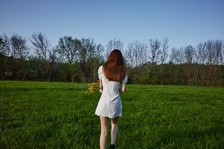 女士穿裙子摄影照片_日落时分，一位身穿白色裙子、留着长红头发的女士站在绿色田野中，背对着镜头