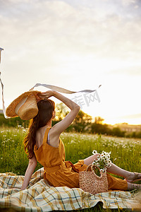 一位身穿橙色裙子的女士坐在洋甘菊花田里，手里拿着一顶草帽，享受着夕阳