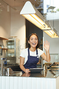 快乐的亚洲女咖啡师在咖啡店工作，向客户挥手，在咖啡馆柜台后面处理订单，穿着蓝色围裙