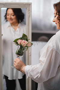 一位身穿白衬衫的成年女性站着，手里拿着洒有玫瑰的玻璃杯。