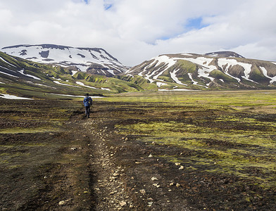 孤独的徒步旅行者在冰岛自然保护区 Fjallabaki 的小径、绿草苔藓草甸和积雪覆盖的流纹岩山上行走