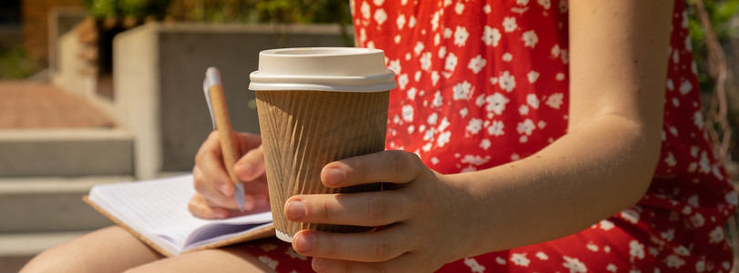 面目全非的年轻女子穿着红色连衣裙，喝着牛皮纸杯里的咖啡，在木凳上写下感恩日记。