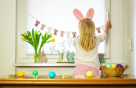 复活节摄影照片_一个穿着粉红色摇滚和白色衬衫的小女孩正在为复活节装饰房间。