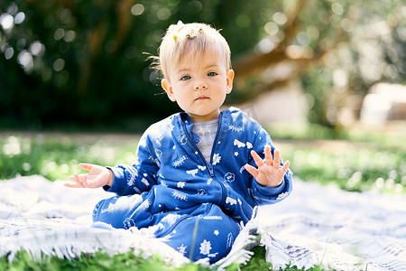 身穿蓝色工装裤的小孩坐在绿色草坪上的毯子上，在绿树背景下展示他的手掌