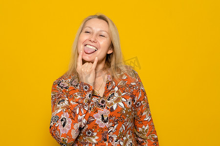 摇滚手势摄影照片_身穿带图案连衣裙的白人金发女子做出摇滚手势，同时以一种无忧无虑、厚颜无耻的态度伸出舌头，在黄色背景中被隔离。