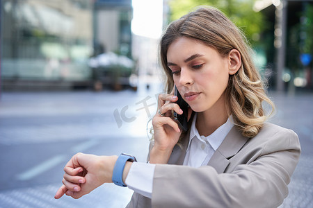 严肃的女商人在手表上看时间，用手机打电话，在城里等人