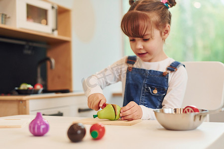 玩玩具的小女孩摄影照片_穿着休闲服的小女孩坐在桌边，在厨房玩玩具玩得开心