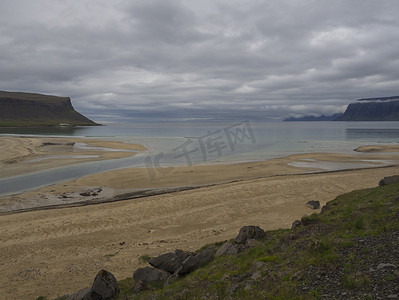 在冰岛西部峡湾的粉红色米色沙滩和绿草和石岸上欣赏绿色悬崖和海洋、河流三角洲和喜怒无常的天空背景