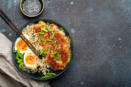 亚洲面条拉面汤、油炸的 panko 鸡片和煮鸡蛋在陶瓷碗里用筷子和酱油在石头质朴的背景顶视图上，文字空间