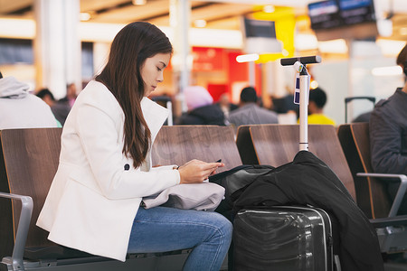 机场女乘客拿着手机等待延误的航班坐在航站楼门口，带着行李。