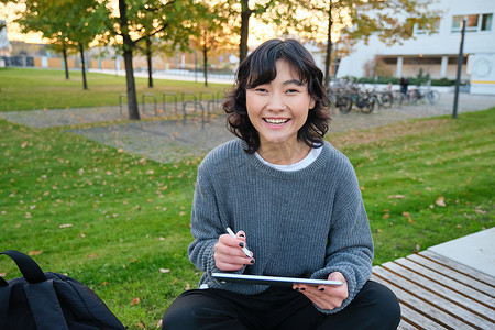 年轻女孩、韩国艺术家或艺术学生坐在公园里，拿着数字平板电脑，用图形笔画画，抓取设计或项目，环顾四周