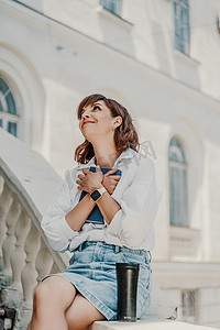 牛摄影照片_一位身穿白衬衫和蓝色牛仔裙的女士坐在栏杆上，将蓝色记事本抱在建筑物上。