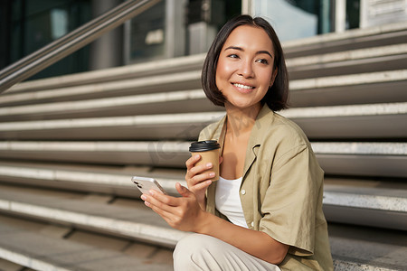 都市女孩摄影照片_快乐的都市女孩喝着她的外卖咖啡，在智能手机上滚动。